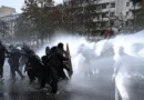 Protesters take cover behind a plywood board as French riot police use a water canon amid clashes during an authorised demonstration organised by labour unions in Paris as part of nationwide protests called by the